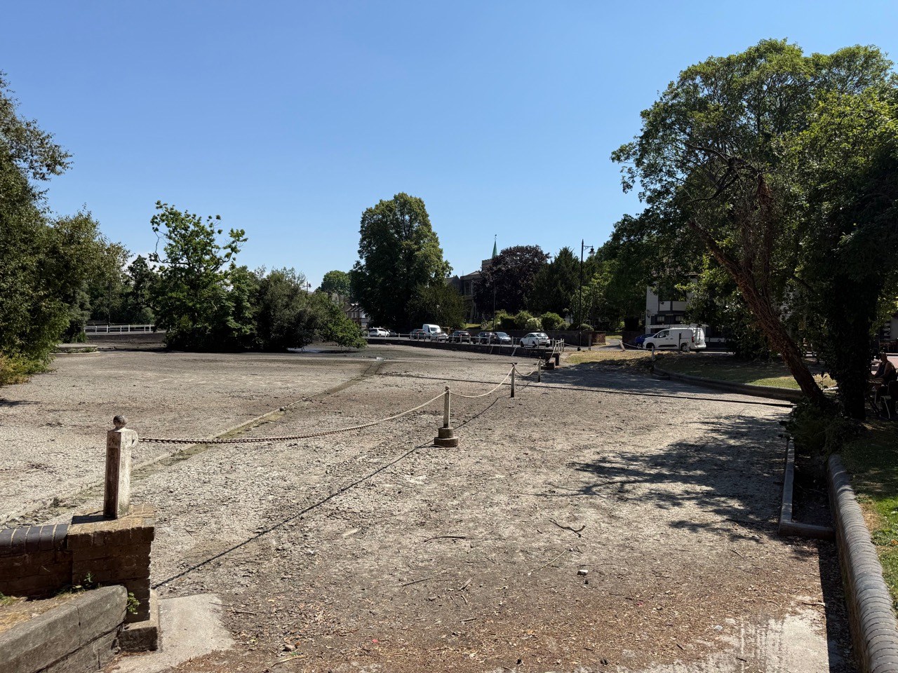 The drained upper pond at Carshalton, showing the exposed pond bed, chains, trees and nearby buildings beneath a clear blue sky
